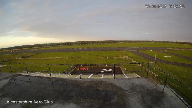 A wide view of the Leicestershire Aero Club grounds during sunset. The scene features a large, grassy area bordered by a wooden fence. In the foreground, there is a marked helipad with an "X" shape on the ground. A paved pathway leads away from the helipad. In the distance, a main runway is visible, alongside various taxiways. The sky is partly cloudy with soft hues of gray and orange as the sun sets.