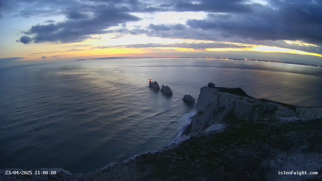 A serene coastal scene during sunset, featuring calm waters reflecting hues of orange and purple in the sky. In the foreground, a rocky cliff descends toward the sea, with patches of greenery. Prominently, a lighthouse stands on a rock formation in the water, its light glowing red. In the distance, faint lights from a shoreline are visible against the twilight sky. The scene conveys tranquility and natural beauty.