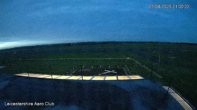The image shows a dark, overcast sky with a bluish hue as evening approaches. In the foreground, there is a structure with a partially illuminated ledge, and below it, a helicopter landing pad marked by a large white cross. To the right, an open green field stretches out, with a distant airport runway visible. The timestamp at the top indicates the date and time as April 23, 2025, 21:00:22. The setting appears quiet with no visible aircraft or people.