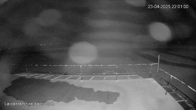 The image shows a dark scene captured by a webcam at the Leicestershire Aero Club. There is a paved surface in the foreground with several rectangular sections marked on it. In the background, the scene appears foggy or obscured, with faint light sources visible in the distance. A railing can be seen on the right side of the image, and the date and time are displayed in the upper right corner, indicating it is night. Overall, the image conveys a dimly lit, atmospheric environment.