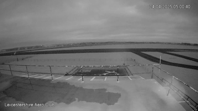 A black-and-white image from a webcam showing the grounds of Leicestershire Aero Club. The scene appears to be early morning with overcast skies. In the foreground, there are parking spaces with signs and a small paved area with a large "X" painted on the ground. Beyond this, there are open fields and some fencing, with a distant horizon and low hills. The timestamp in the top right corner indicates the date as April 24, 2025, and the time as 5:00 AM.
