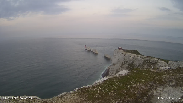 A wide view of a coastal scene with white chalk cliffs meeting the ocean. There are two red and white navigation lighthouses on the cliff edge and a series of white chalk stacks extending into the water. The sky is overcast with grey clouds, and the water appears calm.