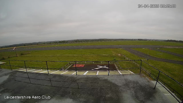 A view from a webcam at LeicesterShire Aero Club showing a runway and grassy field under a cloudy sky. The timestamp on the image reads "24-04-2025 05:59:59." The foreground includes a small platform with a safety railing, and there are marked parking spaces and a helicopter landing pad below. The scene appears to be early in the morning or late in the evening with overcast weather.