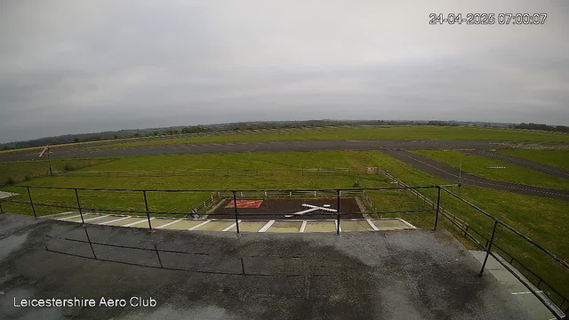 A wide view of an airfield with a grassy runway, taxiways, and open skies overhead. In the foreground, there is a concrete platform with a metal railing, and a small helipad marked with a white helicopter symbol. The timestamp in the upper right corner reads 24-04-2025 07:00:07. The bottom left corner shows the text "Leicestershire Aero Club."