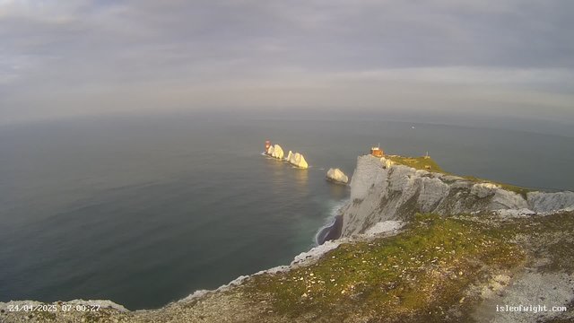 A view of white chalk cliffs rising steeply from the ocean, with large white rock formations offshore. There is a small building and a lighthouse on top of the cliffs, and the sky is cloudy with soft light. The ocean appears calm, with gentle waves near the shore. The image has a timestamp at the bottom left corner and a website URL at the bottom right.