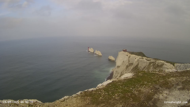 A scenic view of tall white chalk cliffs extending into the ocean, with large rock formations and a lighthouse visible on one of the cliffs. The sky is partly cloudy, and the water appears calm. There is a pathway along the top of the cliffs with a fence, and the image has a timestamp of April 24, 2025, at 8:00:31.