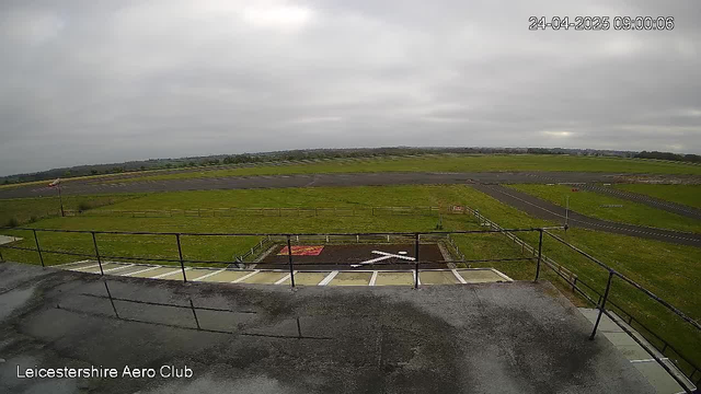An outdoor view from a webcam at Leicestershire Aero Club shows an overcast sky with gray clouds. In the foreground, there is a flat rooftop with a metal railing around the edge. Below the rooftop, there are a few parking spaces and a small fenced area with an arrow painted on the ground pointing to the right. Beyond that, there is a grassy field with a runway and taxiways of the aerodrome stretching into the distance. The landscape is flat, and there are some distant trees and low hills on the horizon. The date and time displayed in the upper right corner are April 24, 2025, at 09:00:06.