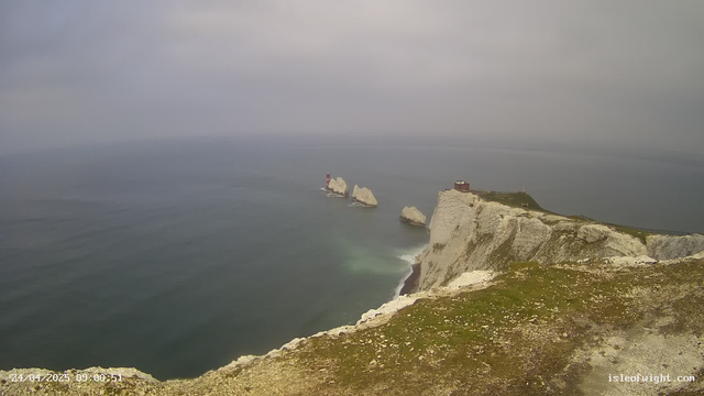 A view of white chalk cliffs overlooking the ocean. Several large rock formations rise from the sea near the cliffs. At the top of the cliff, there is a small building and a lighthouse in the distance, with a fence running along the edge. The sky is overcast, and the water below appears calm. The timestamp at the bottom left reads "24/04/2023 09:00:51," and the bottom right has the text "islotlight.com."