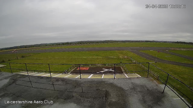 A view of the runway and surrounding grassy area at Leicestershire Aero Club on a cloudy day. The image is taken from an elevated position, possibly a control tower or observation deck, with a weathered roof and railing in the foreground. The runway stretches into the distance under a grey, overcast sky. There are no aircraft visible on the runway or in the sky. The timestamp in the upper right corner indicates the date and time as April 24, 2025, at 10:00:16.