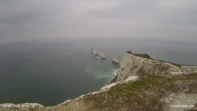 A view from a webcam showing white chalk cliffs on a coast, with a lighthouse and a small building on top of the cliffs. The sea is calm with gentle waves, and the sky is overcast with gray clouds. The scene appears to be during daylight hours.