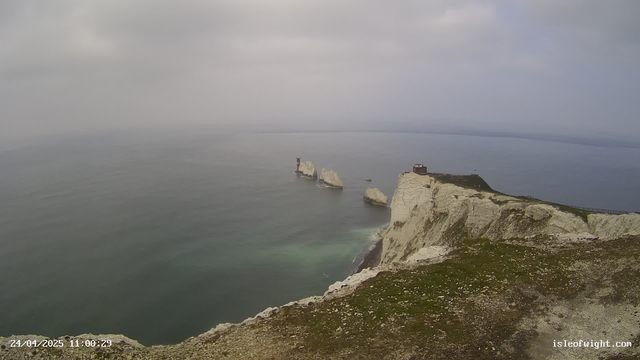 A view of tall white chalk cliffs rising from the ocean, with several large chalk rocks extending into the water near the base of the cliffs. On top of the cliffs, there is a small building and a walking path or railing along the edge. The sky is overcast with gray clouds, and the water appears calm with gentle waves. A timestamp and website URL are visible in the bottom corners.