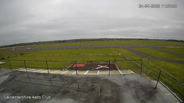 A view from a webcam showing an outdoor area at Leicester Airport. In the foreground, there is a parking lot with a few marked parking spaces and a railing. Beyond the parking lot, a grassy field surrounds a small runway, with a single small aircraft parked on the tarmac. The sky is overcast with gray clouds, and the landscape extends to a distant horizon with some low hills. The timestamp in the top right corner reads 24-04-2025 11:00:58.