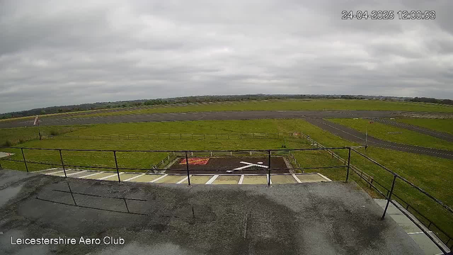 A wide view of an airfield with runways and grassy areas under a cloudy sky. A small helipad with a white helicopter symbol and an arrow marks the landing zone. The modern railing and observation deck are visible in the foreground. The date and time are displayed in the top right corner, and "Leicestershire Aero Club" is written in the bottom left corner.