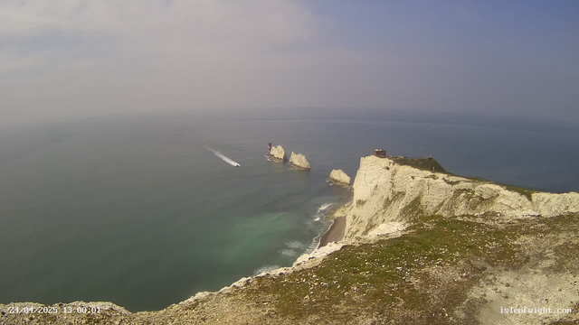 An aerial view of white chalk cliffs along a coastline with green grass on top. There is a building on the far right side of the cliffs. In the water, a boat is leaving a white wake as it moves past large rock formations extending into the sea. The sky is partly cloudy with blue patches. The image is timestamped "24/04/2025 13:00:01" in the bottom left corner.