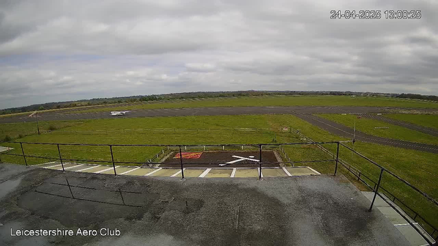 An outdoor view of an airfield on a cloudy day. In the foreground, there is a flat rooftop with a railing and parking spaces marked with white lines. A small aircraft is visible on the tarmac in the middle distance, and the surrounding area is grassy with a few pathways and fences. The sky is mostly cloudy with some patches of blue. The timestamp in the top right corner indicates the date and time as April 24, 2025, 13:00:25, and the bottom left corner shows "Leicestershire Aero Club."