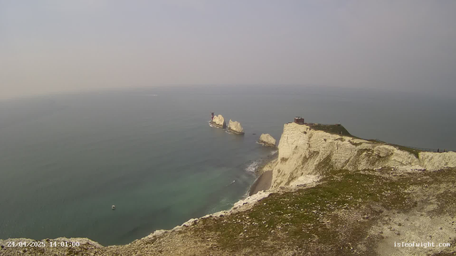 A view of high white chalk cliffs rising from the ocean, with a small building or observation post at the top of the closest cliff. In the distance, more white cliffs and rock formations jut out from the sea. The water below is calm, with a boat visible near the bottom of the image. The sky is overcast with a light gray hue.