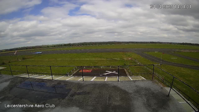 Sky with scattered clouds over an open grassy area. A small blue airplane is taxiing on the runway in the distance. The scene is viewed from a webcam at Leicesterhire Aero Club, with a timestamp of 24-04-2025 at 14:01:01. The foreground shows part of a platform or viewing area with black railings.