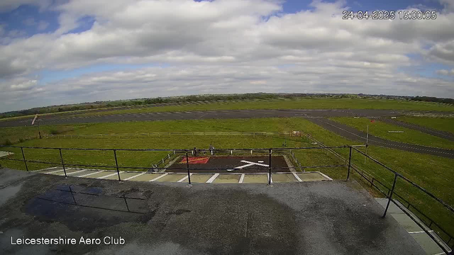 A wide view of an airfield showing a grassy area, a small runway, and a large open sky with scattered clouds. There are some marked areas on the ground, a few small structures, and a fence. The timestamp in the upper right corner indicates the date and time as April 24, 2025, at 15:00:05. The text "Leicestershire Aero Club" is visible in the bottom left corner.