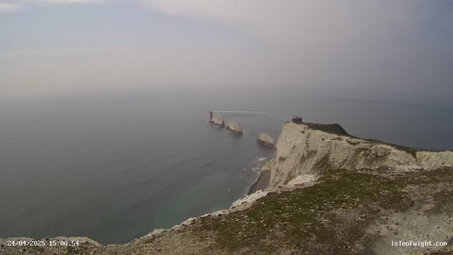 An aerial view of white chalk cliffs along the coastline with a small building or lighthouse at the edge. Several large rocks are visible in the water near the cliffs, and the sea appears calm. The sky is mostly cloudy with soft, diffused light. The image includes a timestamp and the website "isleofwight.com" in the bottom corners.