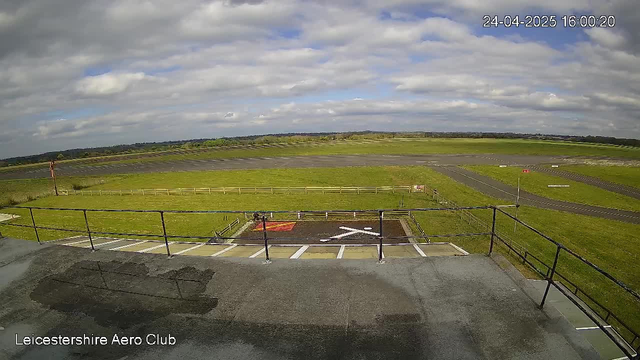 A view from a webcam shows an open grassy area with a few small structures and a paved runway extending into the distance under a partly cloudy sky. The timestamp on the image is 24-04-2025 16:00:20, and the location is indicated as Leicester Shire Aero Club.