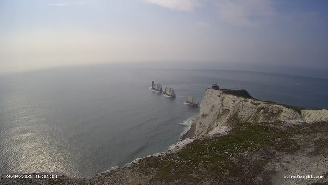 A view of white chalk cliffs rising from the sea, with a grassy top and a building near the edge. Several large rock stacks are visible offshore, and a lighthouse stands on one of the stacks. The sea is calm, with gentle waves, and a sailboat can be seen in the distance. The sky is partly cloudy with soft light.
