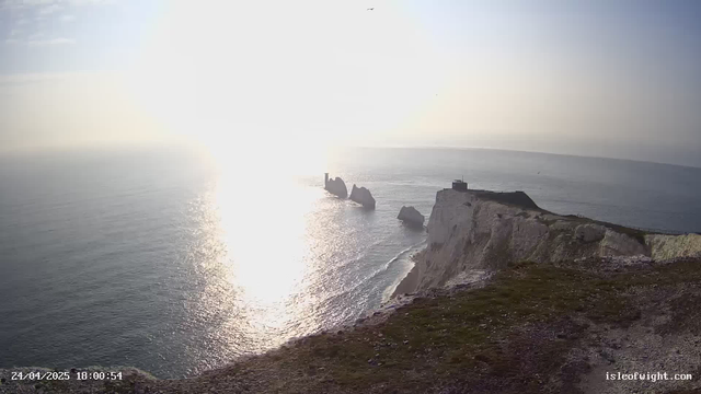 A panoramic view of a coastal landscape featuring white chalk cliffs with rocky formations extending into the water, topped with grassy areas and a small building or lighthouse. The ocean is calm, with sunlight reflecting off its surface, creating a bright glare near the horizon. The sky is mostly clear with a few scattered clouds. The image has a timestamp in the bottom left corner reading "24/04/2025 18:00:54" and a website credit "isleofwight.com" in the bottom right corner.
