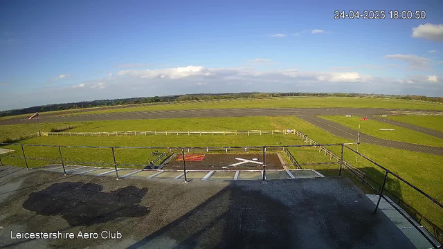 A view from a webcam at Leicesterhire Aero Club showing a clear, sunny day with a bright blue sky and a few scattered clouds. The image captures the airfield's grassy areas and runways, with some small white markers and fencing visible. The timestamp in the upper right corner reads "24-04-2025 18:00:50." The bottom left corner displays the text "Leicestershire Aero Club."