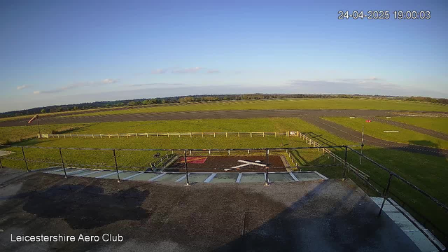 An outdoor view from a webcam at Leicestershire Aero Club showing a clear sky with a few clouds, a large grassy field, and a paved road or runway in the distance. The timestamp in the top right corner indicates the date and time as 24-04-2025 at 19:00:03. The foreground includes a balcony or platform with railings and some equipment or structures near the edge.