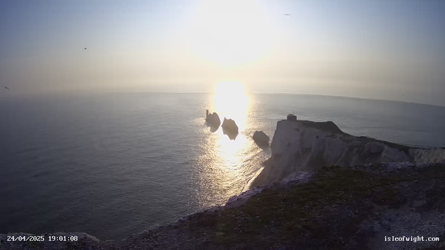 A view of the ocean at sunset with the sun low on the horizon, casting bright reflections on the water. In the foreground, there are white chalk cliffs with a small structure or building near the edge, and several large rocks jutting out of the sea. Several seabirds are flying in the sky above the water. The timestamp in the bottom left corner indicates the date and time as April 24, 2025, at 19:01:08.