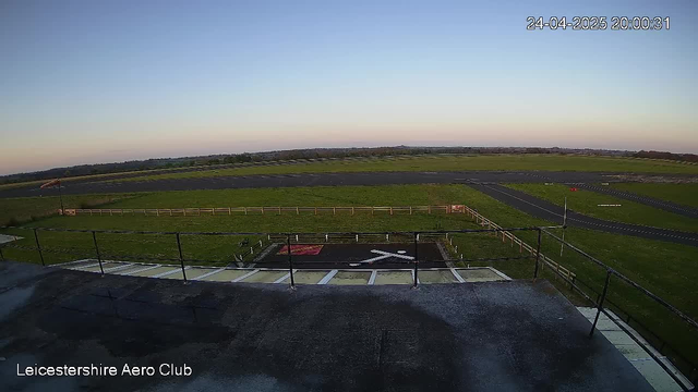 A view from a webcam at Leicestershire Aero Club shows a clear sky with the horizon in the distance. In the foreground, there is a tarmac area with white painted lines and a red and black marking, possibly a helipad or aircraft parking area, surrounded by safety barriers. The surrounding landscape features flat, open fields with a few small trees and some infrastructure such as roads. The timestamp reads 24-04-2025 20:00:31.