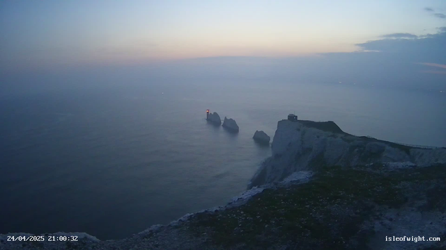 A view of white chalk cliffs overlooking the ocean during dusk or early evening, with a lighthouse on a small rock formation and a taller lighthouse on the cliff. The sky is partly cloudy with soft pastel colors. The date and time at the bottom left corner indicate April 24, 2025, at 21:00:32. The bottom right corner shows the website isleofwight.com.