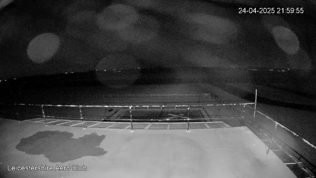 A black-and-white image from a webcam shows an outdoor scene at night with a large empty area surrounded by a metal fence. There is a small, irregularly shaped patch of shadow on the ground near the bottom left corner. In the background, faint lights can be seen along the horizon. The top right corner displays the date and time as "24-04-2025 21:59:55," and the bottom left corner has the text "Leicestershire Aero Club." The image appears to be taken at an airport or airfield, possibly a runway or taxiway, with minimal visible activity.