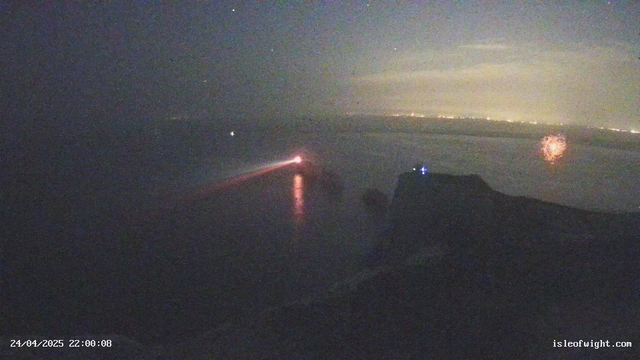 A nighttime scene captured from a webcam shows a dark expanse with faint reflections on a body of water. There is a red navigation light on the left side, casting a glow on the water’s surface. To the right, a silhouette of a structure, possibly part of an aircraft or boat, is visible with two small blue lights. In the background, faint city lights stretch across the horizon, illuminating the distant shoreline. The image has a timestamp of "24/04/2025 22:00:08" and a website link at the bottom right corner reading "isleofwight.com".