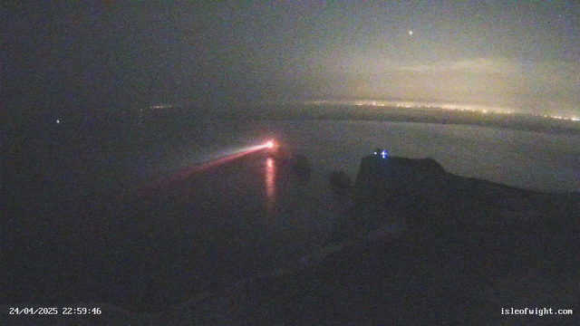 A view from an airplane at night showing the dark ocean below with faint reflections of red and white navigation lights on the water. The horizon displays a distant cityscape with twinkling lights under a starry sky. The airplane wing is partially visible on the right side of the image.
