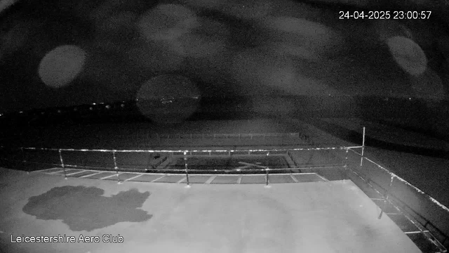 A black and white nighttime image from a webcam shows an empty outdoor area with a small, irregularly shaped patch of shadow or puddle on the ground in the foreground. There is a railing or barrier lining the edge of the area, which appears to be a viewing or sports platform. The background consists of a dark, cloudy sky with a few faint lights visible in the distance. The timestamp in the top right corner reads "24-04-2025 23:00:57," and the bottom left corner has the text "Leicestershire Aero Club."