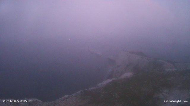 A view of a lighthouse on a dark, rocky shoreline at dawn or dusk, with a hazy sky and an ocean in the background. The timestamp in the bottom left corner reads "25/04/2025 04:59:49," and the bottom right corner has the website "isleofwight.com."