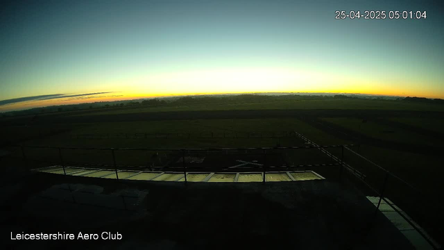A wide view of the early morning sky at sunrise over a flat, open landscape with fields and sparse trees, captured from a webcam at Leicestershire Aero Club. The sky transitions from dark blue to a soft yellow near the horizon. The timestamp in the upper right corner indicates the date as April 25, 2025, and the time as 05:01:04. The bottom left corner features the label "Leicestershire Aero Club."