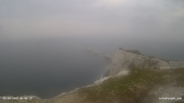 A foggy seascape featuring towering white chalk cliffs with patches of green grass on top. A small building or structure is visible on the edge of the cliffs. The sea below is calm and partially obscured by fog, creating a mysterious and tranquil atmosphere. The image includes a timestamp of "25/04/2025 06:00:37" and a website credit "isleofwight.com" in the lower corners.