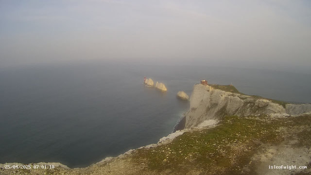 A scenic view of white chalk cliffs overlooking the ocean, with several large rocks and stacks standing in the water near the cliffs. The sky is overcast, and the photo appears to be taken early in the morning. There are small patches of grass and dirt on the foreground, and a railing along the top of the cliffs. The timestamp indicates the photo was taken on April 25, 2025, at 7:01 AM.