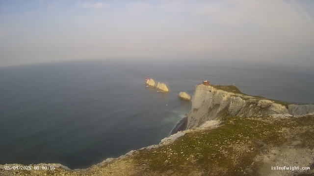 A view of white chalk cliffs dropping into the ocean, with several rock formations jutting out of the water. A small lighthouse is visible on one of the rocks in the distance, and a building sits atop the cliff. The sky is partly cloudy with a soft, diffused light illuminating the scene.