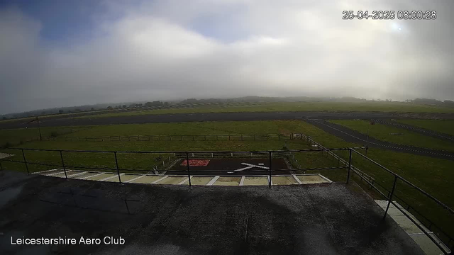 The image shows an outdoor view from a webcam at Leicestershire Aero Club. In the foreground, there is a small helipad with a white helicopter symbol and a red marking. The helipad is enclosed by a black metal railing, and there are a few parking spaces visible at the bottom. Beyond the helipad, there are green grassy fields stretching into the distance, with a winding paved road running through the landscape. The sky is partly cloudy with patches of bright sunlight breaking through, creating a mix of light and shadow across the scene. The timestamp indicates the photo was taken on April 25, 2025, at 8:00 AM.