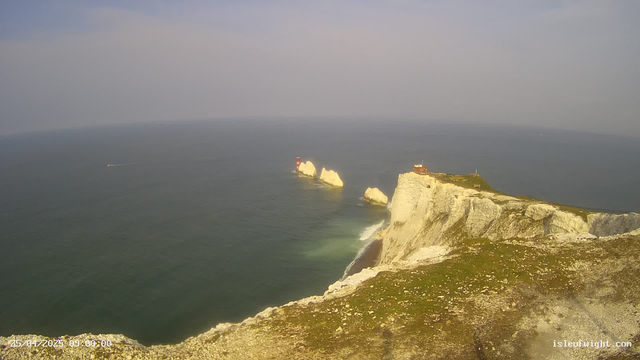 A scenic view of white chalk cliffs extending into the ocean, with a small building or lighthouse at the top of the closest cliff. In the background, the sea stretches to the horizon, with a boat passing by, leaving a wake behind. The sky is mostly clear with a slight haze. The image is timestamped April 25, 2025, at 9:00 AM.