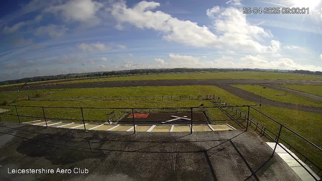 A view of an open field under a partly cloudy sky, taken from a webcam at Leicesterhire Aero Club. The image shows grassy areas, a paved track or runway, some fencing, and a parking area with marked spaces in the foreground. The timestamp indicates it was captured on April 25, 2025, at 9:00 AM.