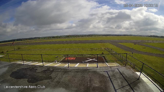 A wide view of a grassy airfield with runways stretching into the distance under a partly cloudy sky. The photo is taken from an elevated platform with a railing, and there are markings for aircraft parking spots in the foreground. The label "Leicestershire Aero Club" is visible in the bottom left corner, and the date and time displayed in the top right corner is "25-04-2025 10:00:15."