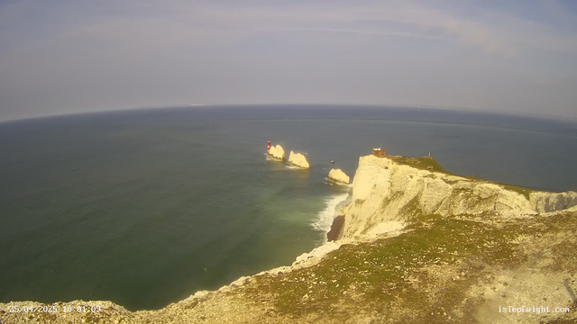 A view of white chalk cliffs extending into the ocean with several large rocks near the shoreline. There is a small building and a lighthouse on top of the cliffs, with a cloudy sky overhead.