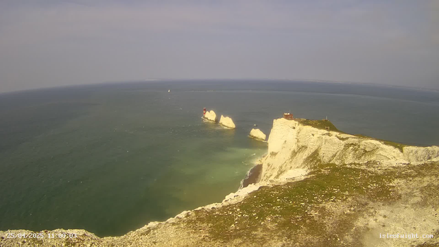 An aerial view of white chalk cliffs along the coast, with a grassy top and a small building on the edge. The sea below is a greenish hue, and several large rock formations are seen in the water extending from the cliffs. The sky appears partly cloudy with a mix of light and darker clouds.