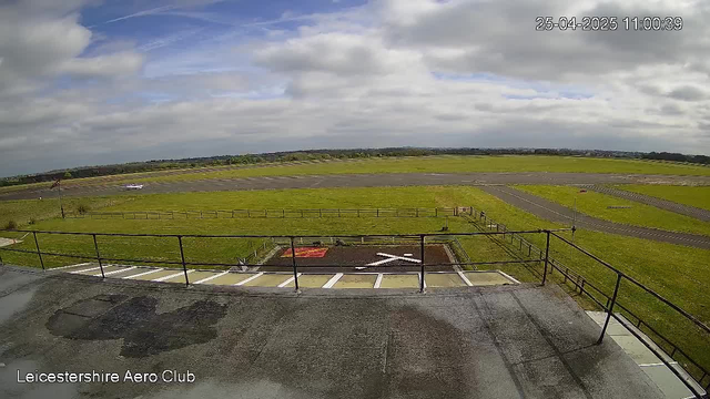 A view from a webcam at Leicesterhire Aero Club showing a runway with an overcast sky. The scene includes a paved area in the foreground with several parking spaces, a grassy field stretching out towards the horizon, and various small structures and equipment along the runway. The timestamp in the top right corner reads 25-04-2025 11:00:39.
