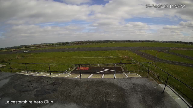 A view from a webcam at Leicesterhire Aero Club showing a runway and grassy airfield under a partly cloudy sky. There are a few small aircraft visible in the distance and parking spaces in the foreground. The timestamp in the top right corner indicates the date and time as April 25, 2025, at 11:59:54.