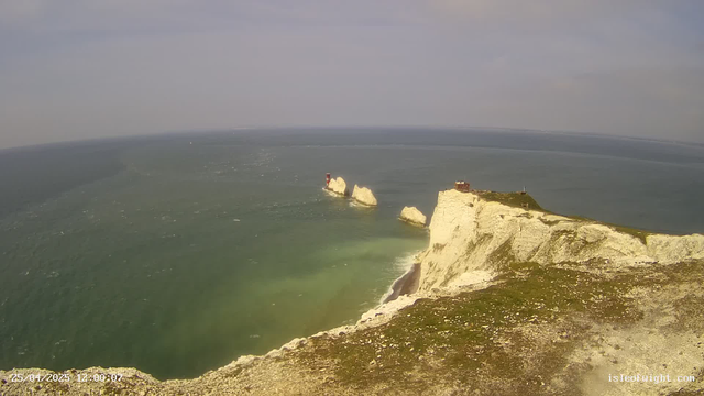 Aerial view of white chalk cliffs along the coastline with a building on top, overlooking the ocean. Several large rocks are visible in the water near the shoreline, and a lighthouse is situated on one of the rocks. The sea is a mix of green and blue hues, with waves crashing against the cliffs. The sky is partly cloudy. The timestamp and website are displayed at the bottom left and right corners respectively.