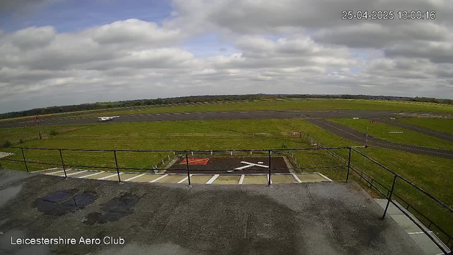 A view of an airport runway with a small airplane in the distance, under a cloudy sky. The image shows a parking area with a red and black marking, white directional arrows, and a railing in the foreground. The bottom left corner has the text "Leicestershire Aero Club," and the top right corner displays the date and time as "25-04-2025 13:00:16."